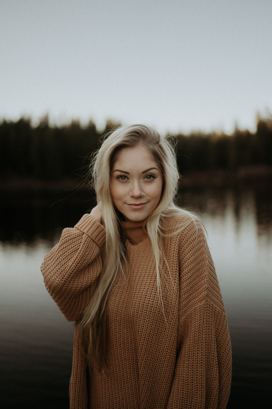 A woman in a brown sweater photographed beside a still lake