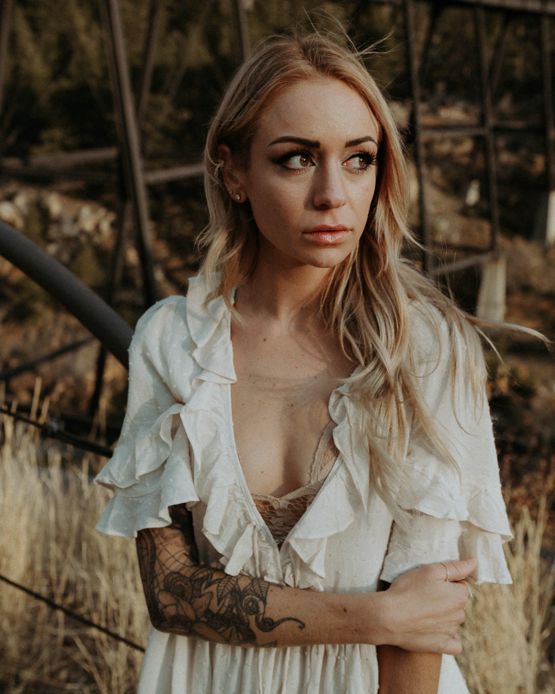 A woman with tattooed arms in a white blouse beside a bridge