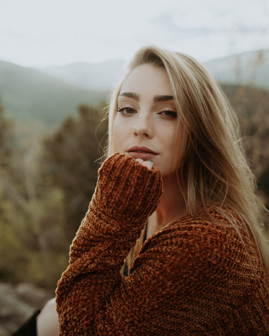A close portrait of a woman in a rust-colored sweater with mountains behind her