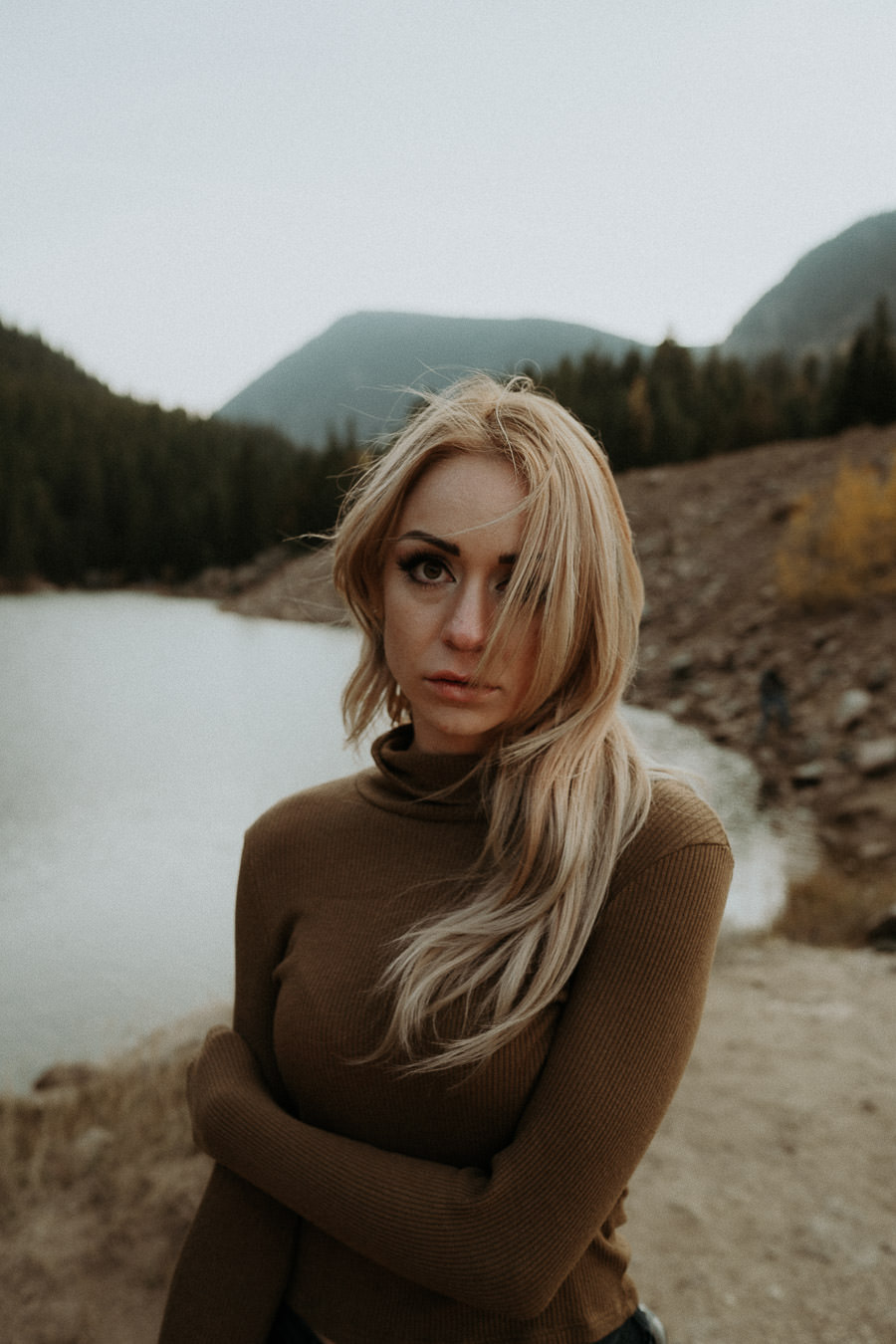 A woman in a brown top looking toward the camera beside a lake