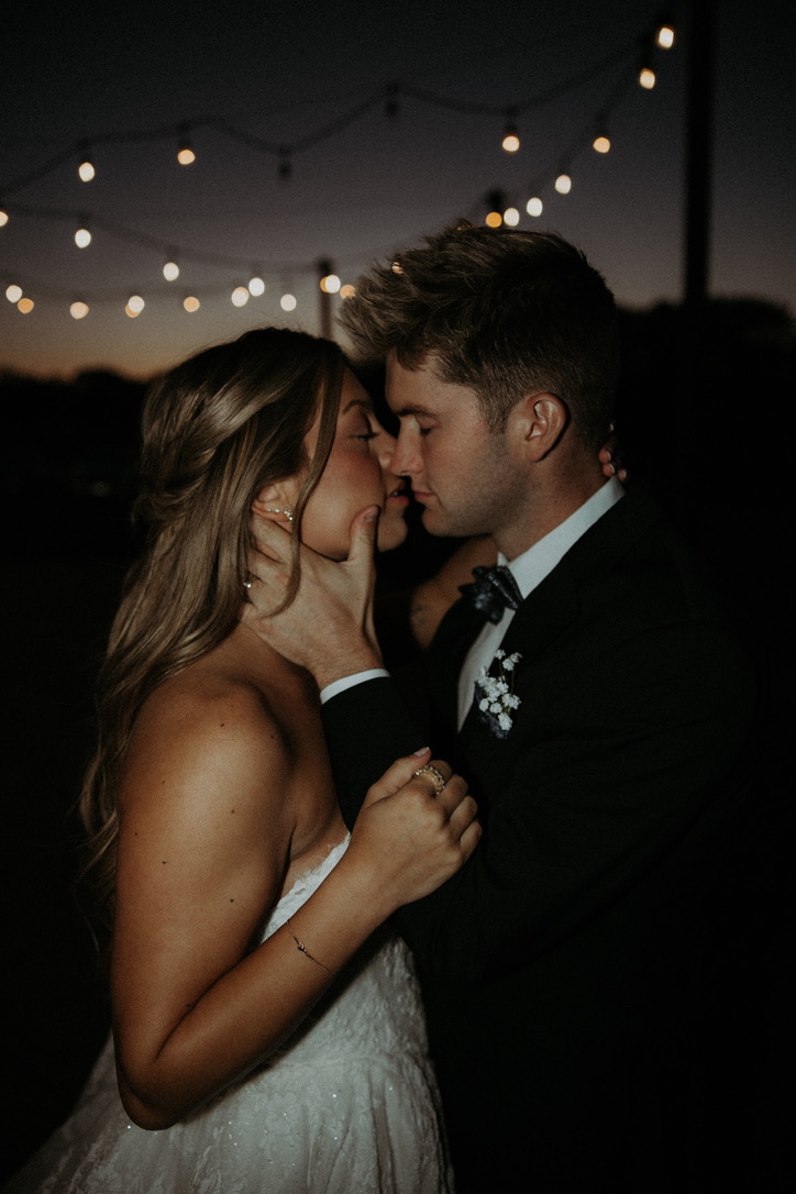 Newlyweds leaning in for a kiss beneath string lights at dusk